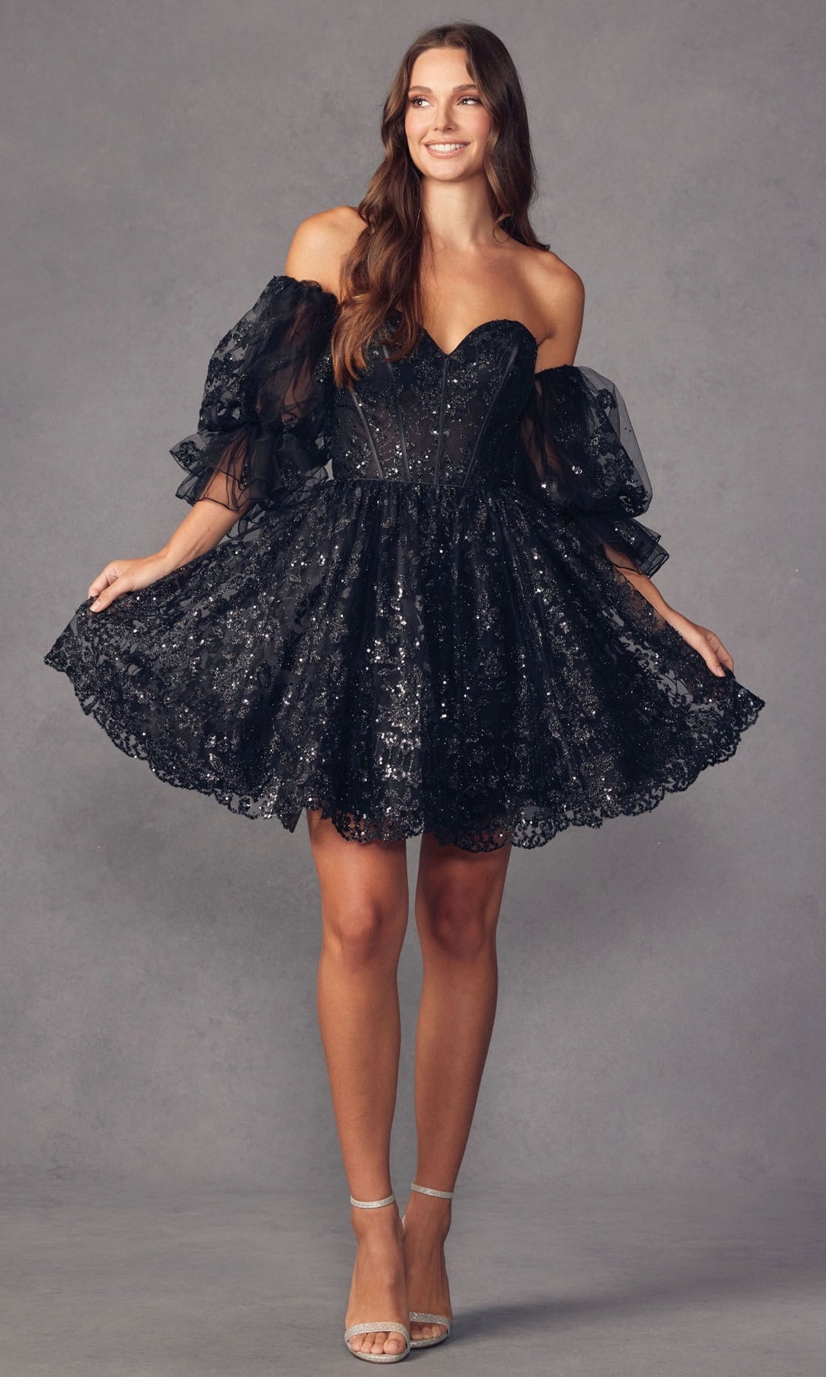 A woman in the Puff-Sleeve Short Strapless A-Line Prom Dress 895—black lace, short length, off-the-shoulder with removable puff sleeves—pairs it with silver high-heeled sandals and poses against a plain gray background.